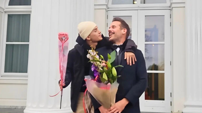 Lars and Sebas celebrating their partnership, smiling warmly at each other while holding flowers outside a white building.