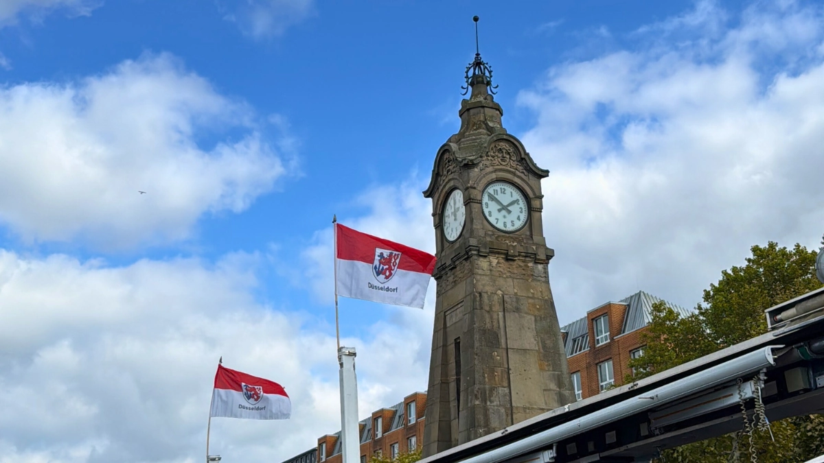 Clock tower with Düsseldorf city flags waving under a blue sky, marking the start of a new chapter in Lars’ journey.